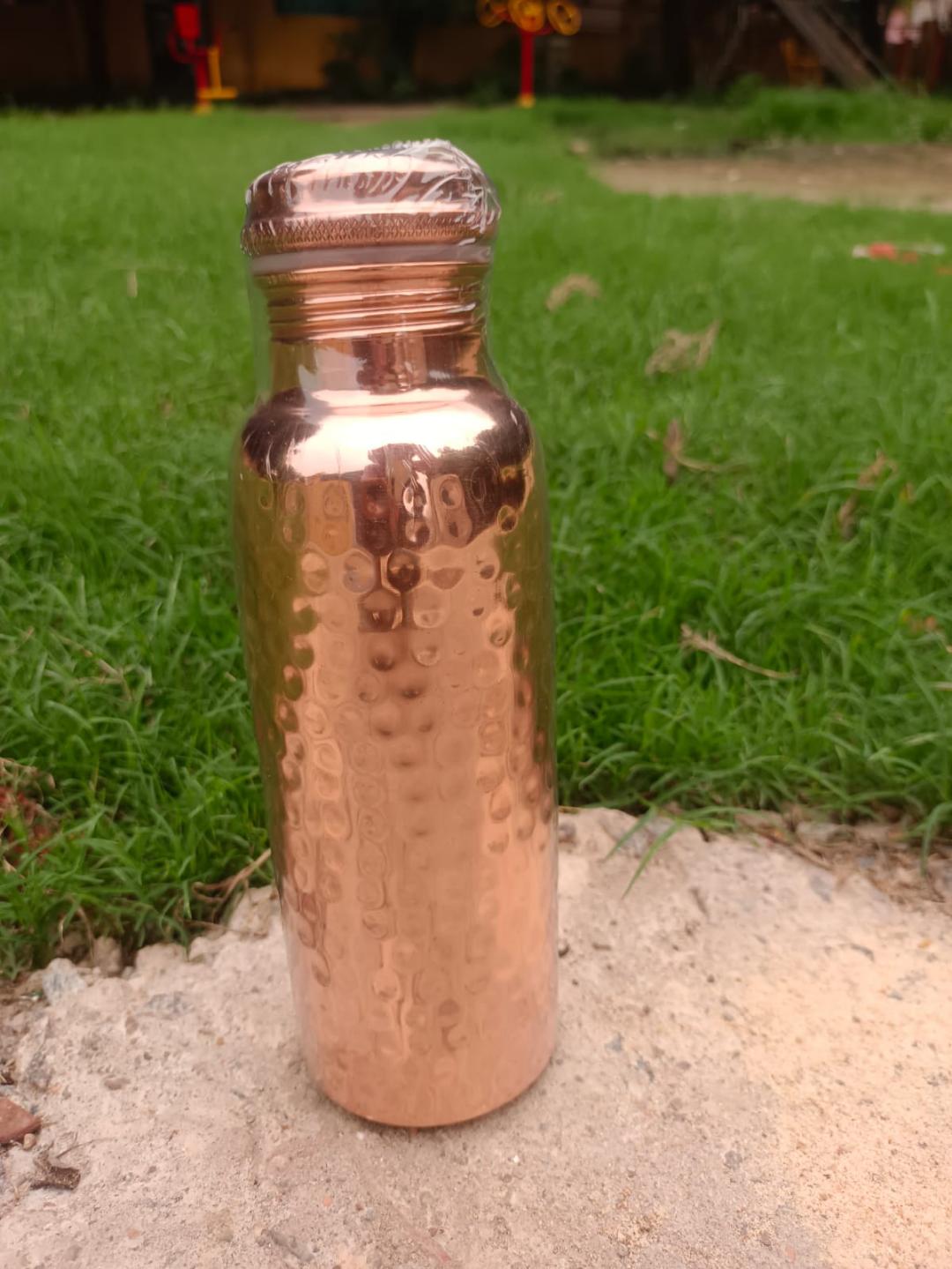A hammered copper water bottle with a textured metallic finish and a crimped screw cap, standing on a sandy surface with green grass in the background.