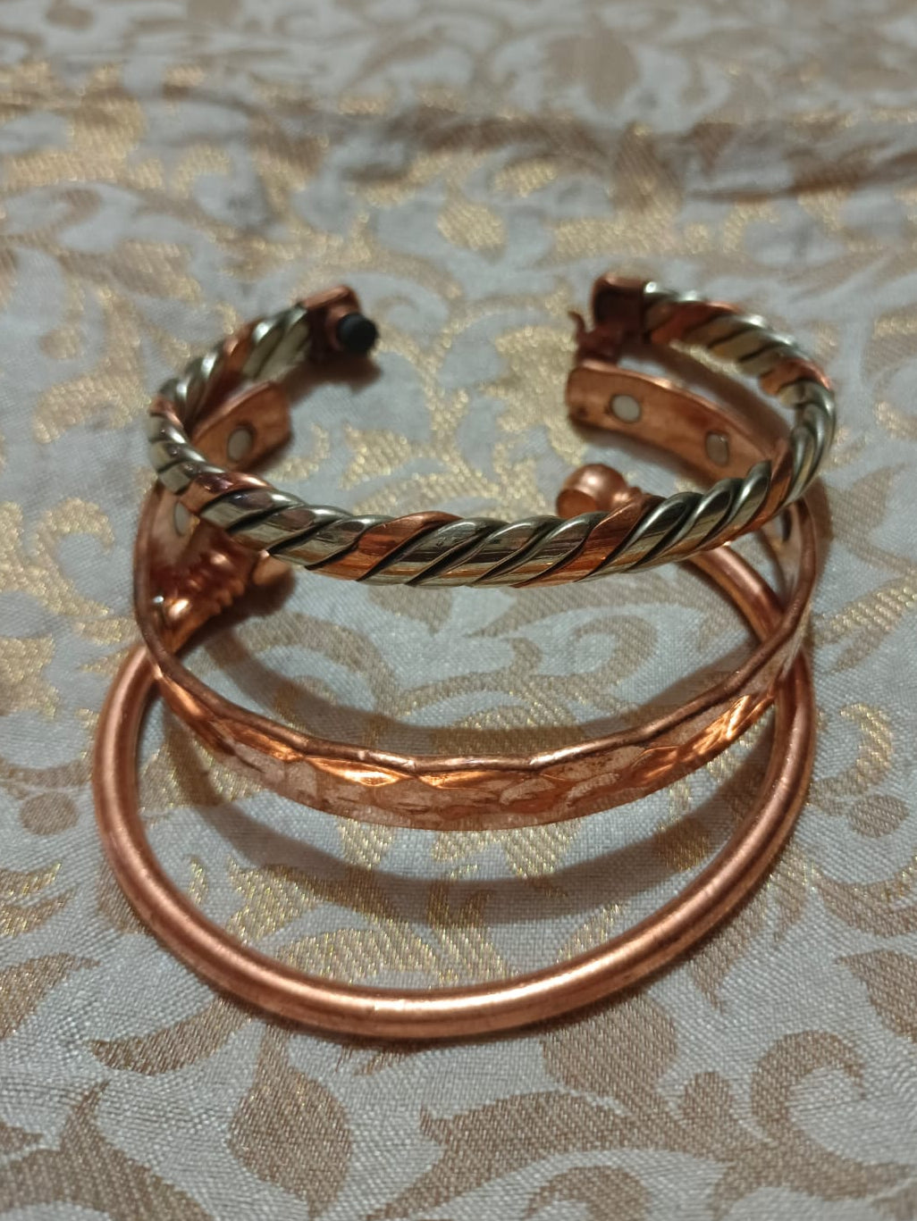 Three copper-toned twisted and smooth bangles stacked together on a beige and gold patterned fabric background.